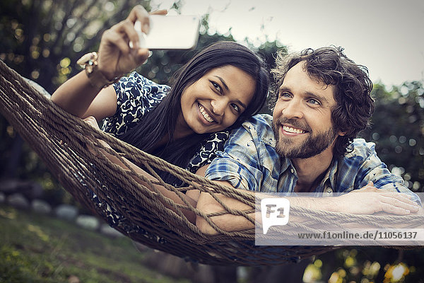 Ein Ehepaar  ein junger Mann und eine junge Frau  die in einer großen Hängematte im Garten liegen und sich ein Selfy von sich selbst nehmen.