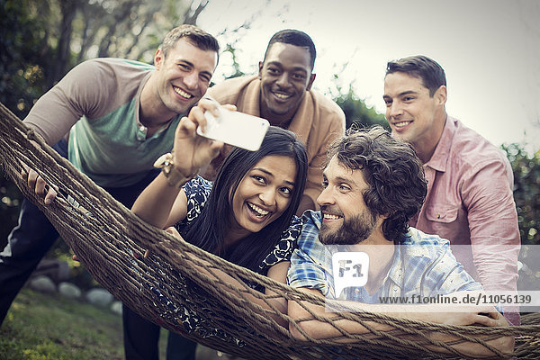 A group of friends lounging in a large hammock in the garden having a beer  and taking a selfie.