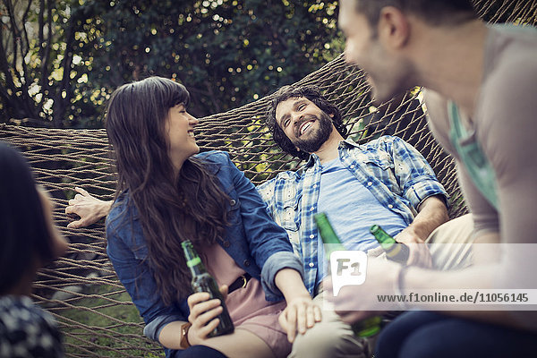 Eine Gruppe von Freunden faulenzt in einer großen Hängematte im Garten bei einem Bier