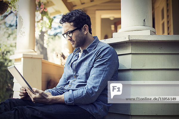 A man sitting relaxing in a quiet corner of a porch  using a digital tablet.