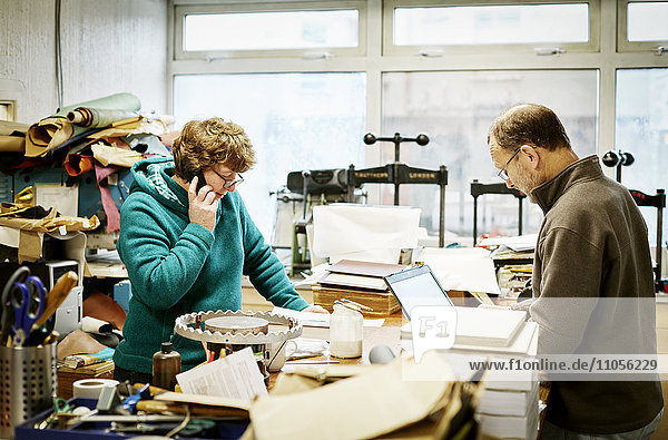 A woman taking a phone call and a man working on a laptop computer in a bookbinding workshop.