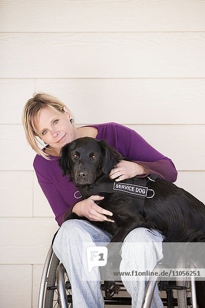 A mature woman wheelchair user with her arms around her service dog  a black Labrador whose front paws are on her lap.