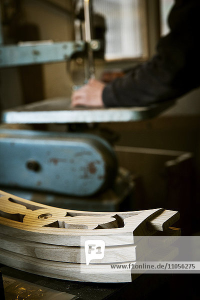 Ein Mann  der in der Werkstatt eines Möbelherstellers mit einer mechanischen Säge arbeitet. Hölzerne Stuhllehnen auf einer Werkbank gestapelt.