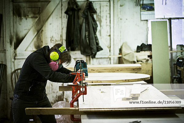 Eine Frau  die in der Werkstatt eines Möbelherstellers arbeitet und mit einer Säge Holz schneidet.
