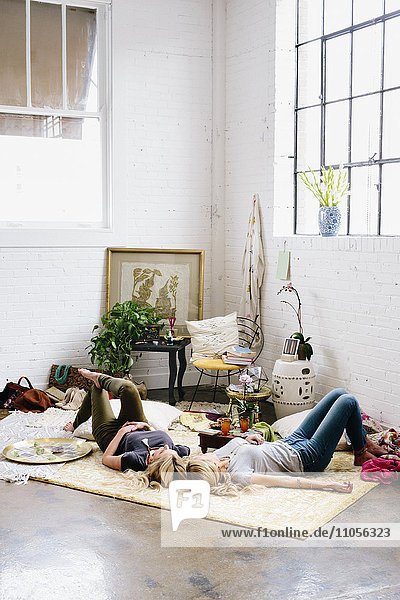 Zwei Frauen liegen auf dem Boden eines mit Kissen und persönlichen Gegenständen übersäten Raumes.