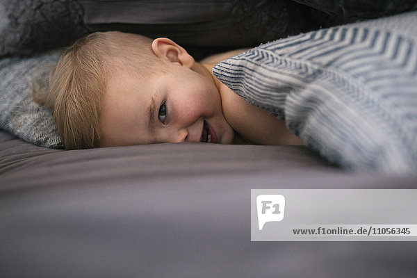 Ein kleiner Junge liegt auf dem Bauch im Bett  schaut in die Kamera und lacht.