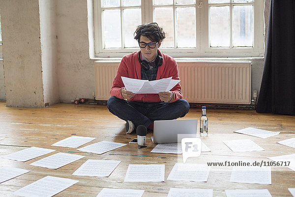 Junger Mann sitzt in einem Probenstudio auf dem Boden  benutzt einen Laptop-Computer und schaut sich Notenblätter an.