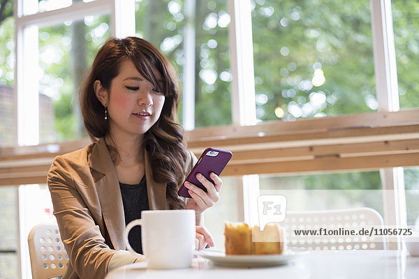 Smiling young woman sitting at a table with a mug and slice of cake  looking at her smart phone.