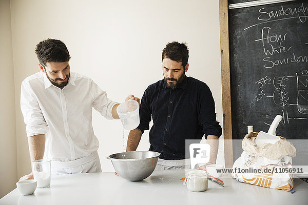 Two bakers standing at a table  preparing bread dough  baking ingredients and a blackboard on the wall.