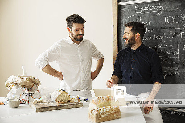 Zwei Bäcker  die mit einem Laptop an einem Tisch stehen  frisch gebackenes Brot  eine Tafel an der Wand.
