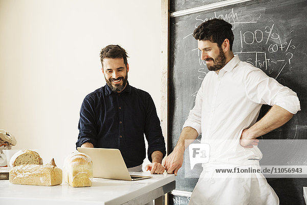 Two bakers standing at a table  using a laptop computer  freshly baked bread  a blackboard on the wall.