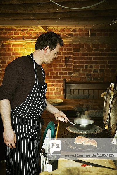 A man in a blue and white striped apron cooking fish on a hot plate on a stove.