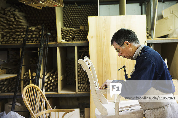 Man standing in a carpentry workshop  working on a wooden chair marking the armrest joint with a pencil.