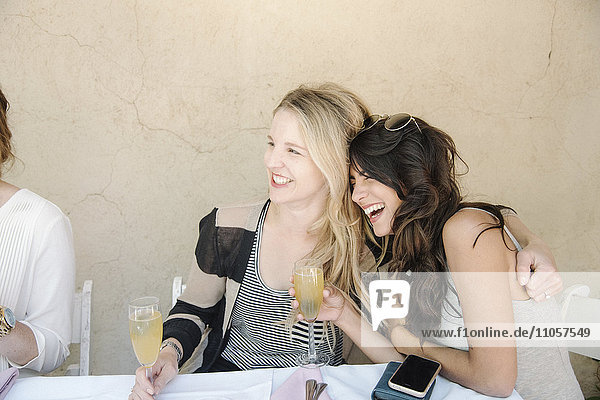 Zwei Frauen sitzen an einem Tisch  umarmen sich und lachen  halten Gläser mit Champagner in der Hand.
