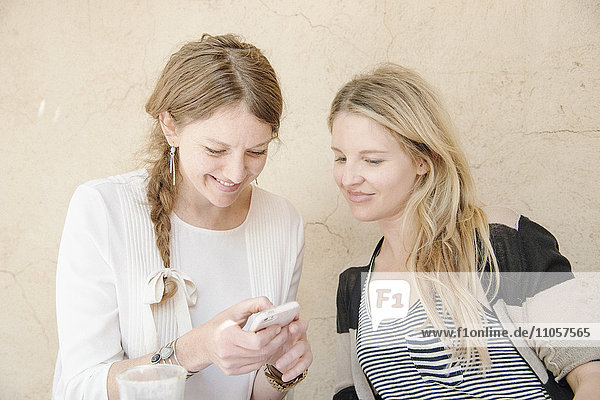 Two smiling women with long blond hair sitting at a table  looking at a cell phone.