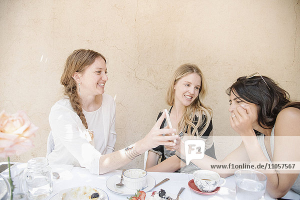 Drei lächelnde junge Frauen sitzen bei Speis und Trank um einen Tisch und schauen auf ein Handy.