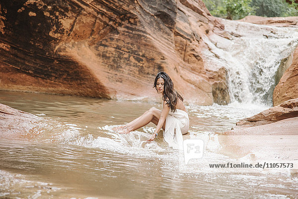 Junge Frau mit langen braunen Haaren  trägt ein langes weißes Kleid und sitzt auf einem Felsen in einem Fluss.