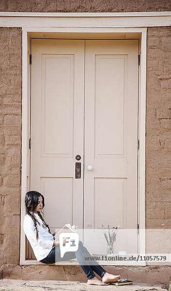 Junge Frau mit langen braunen Haaren  die auf dem Boden vor der Eingangstür eines Gebäudes sitzt.