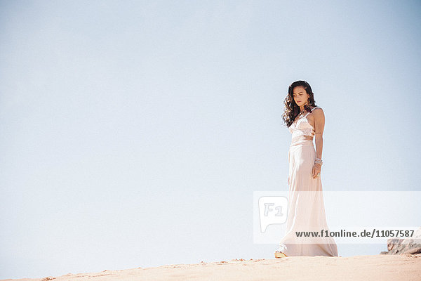 Junge Frau mit langen braunen Haaren  die ein langes weißes Kleid trägt und in einer Wüste steht.