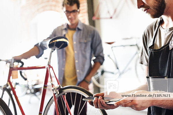 Zwei Männer in einer Fahrradwerkstatt  die sich ein Fahrrad anschauen. Einer hält ein Smartphone und eine Karte in der Hand  die kontaktlose Zahlungen mit einem Smartphone verarbeitet.
