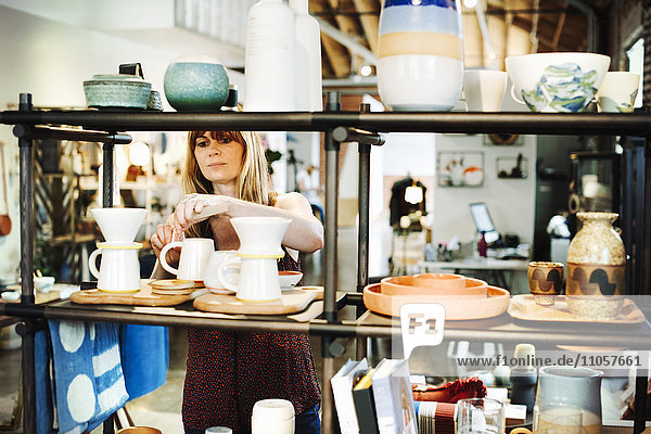 Frau  die in einem Geschäft steht und Keramikobjekte auf einem Regal arrangiert.