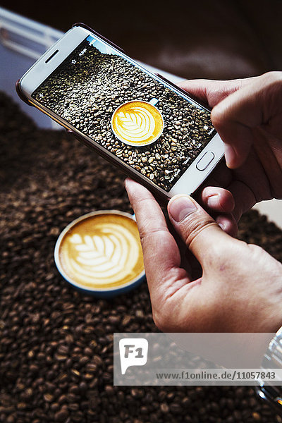 Specialist coffee shop. A person taking a picture with a smart phone of a cup of coffee on top of a heap of roasted coffee beans.