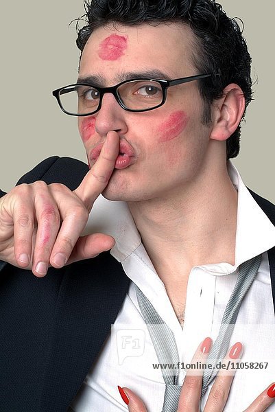 Junger Mann mit Lippenstiftabdrücken im Gesicht Schweigegeste Diskretion