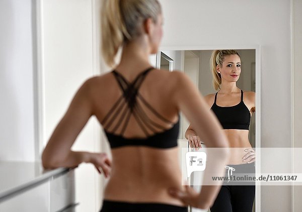 Junge Frau mit Sport-BH und Jogginghose betrachtet sich im Spiegel