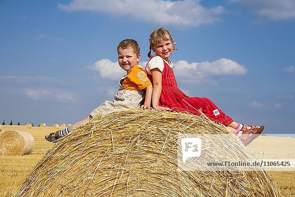 Mädchen und Junge  Geschwister sitzen auf einem Strohballen auf einem abgeernteten Getreidefeld  Deutschland  Europa