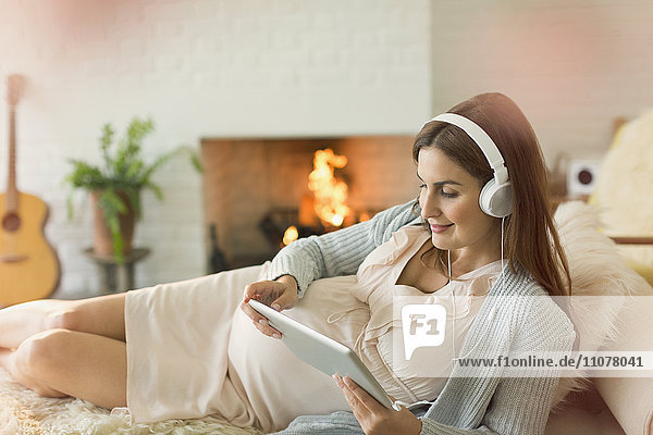 Pregnant woman with digital tablet and headphones listening to music near fireplace