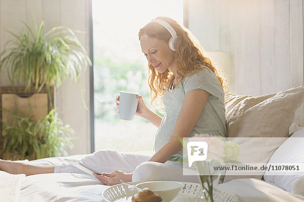 Pregnant woman drinking coffee and listening to music with headphones and digital tablet on bed