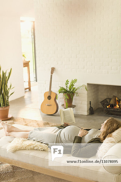 Comfortable pregnant woman laying reading book near fireplace in living room