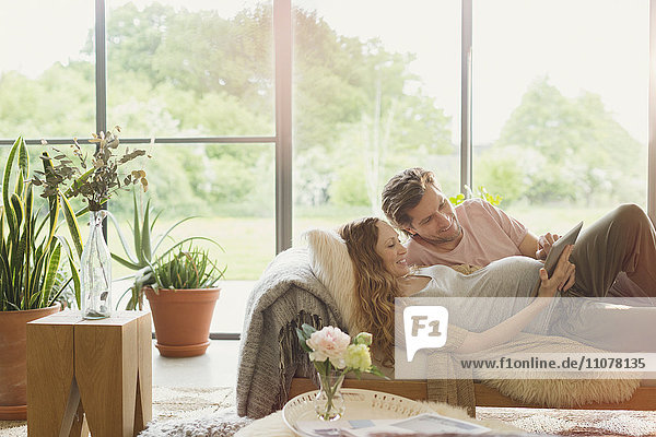 Pregnant couple laying using digital tablet in living room