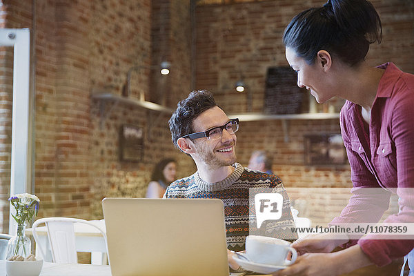 Barista serving cappuccino to smiling man at laptop in cafe