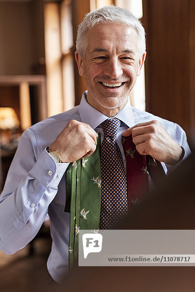 Lächelnder Geschäftsmann beim Anprobieren von Krawatten im Spiegel im Herrenausstattergeschäft