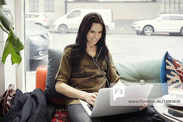 Lächelnde Geschäftsfrau beim Sitzen auf dem Sofa am Fenster mit Laptop