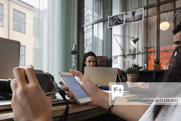 Nahaufnahme eines Bloggers  der Kamera und Smartphone hält  während er am Tisch im Büro sitzt.