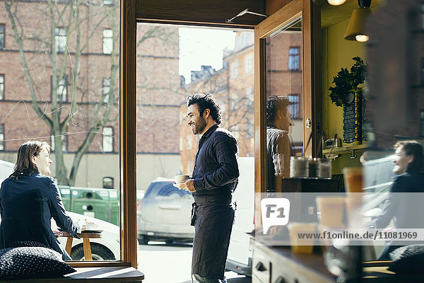 Smiling waiter talking with woman on sidewalk seen through glass at cafe