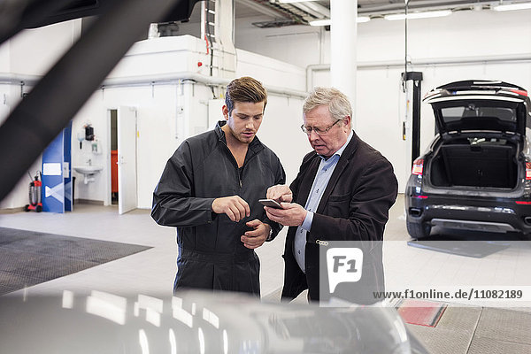 Männlicher Kunde zeigt Handy dem Mechaniker in der Autowerkstatt