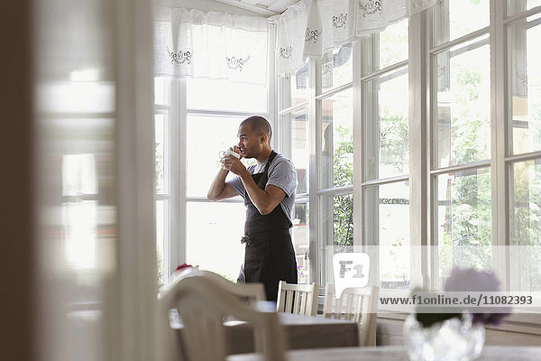 Männlicher Besitzer beim Kaffeetrinken und Telefonieren  während er am Fenster steht.