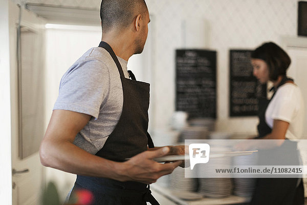 Mann hält Serviertablett mit Blick auf eine Kollegin  die neben einem Tellerstapel im Café steht.