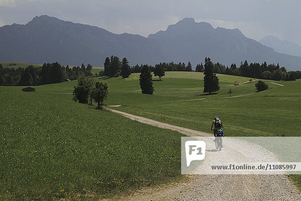 Rear view of person riding bicycle on dirt road amidst grassy field against mountains