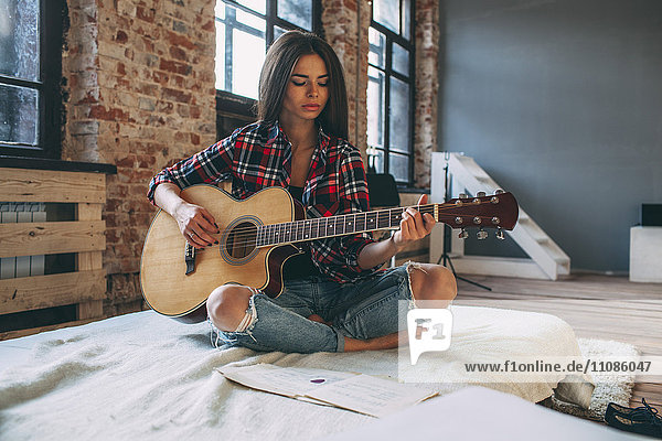 Young woman playing guitar while sitting on bed at home