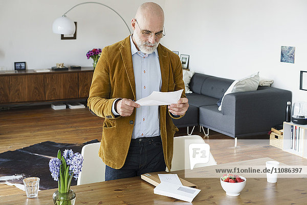 Mature man standing by table and reading letter at home