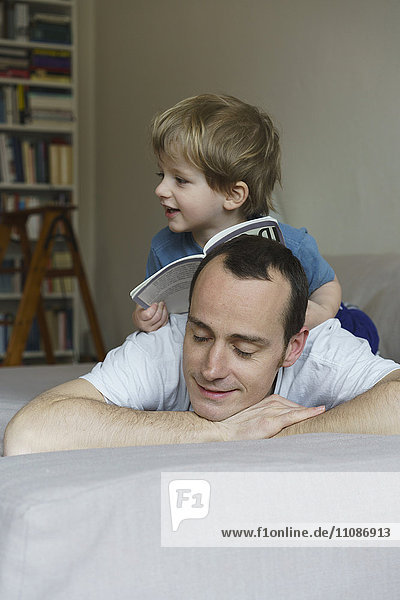 Boy with book sitting on father's back in bedroom