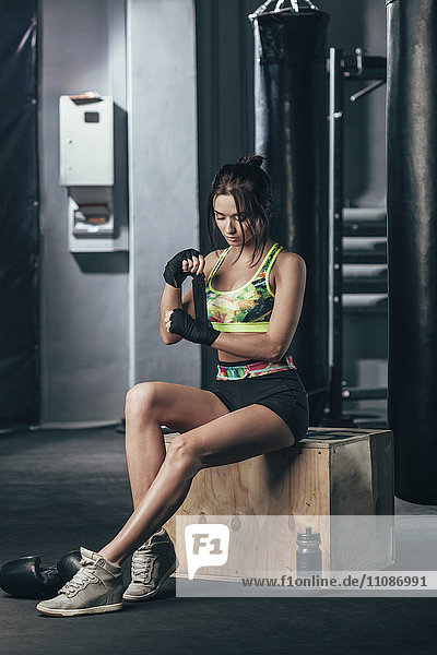 Young female boxer wrapping hand wrap while sitting at gym