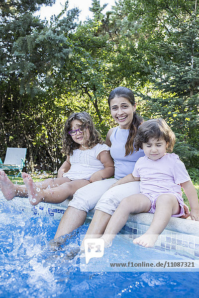 Portrait einer lächelnden Mutter  die mit ihren kleinen Töchtern am Poolrand sitzt.