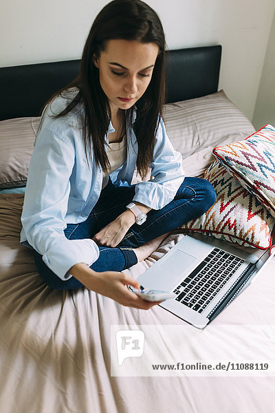 Junge Frau mit Smartphone und Laptop im Bett