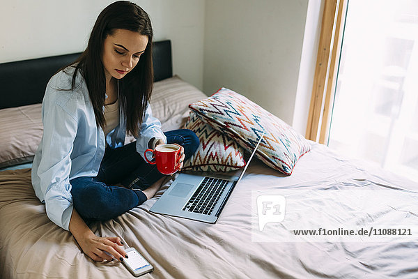 Junge Frau sitzt auf Bett  Kaffeetasse  Laptop und Smartphone