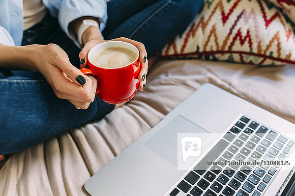 Junge Frau sitzt auf Bett Kaffeetasse Laptop und Smartphone